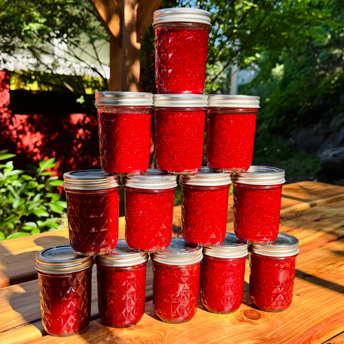 A pyramid arrangement of twelve glass jars filled with bright red jam or preserves sits on a wooden table outdoors under a pergola with green foliage in the background.